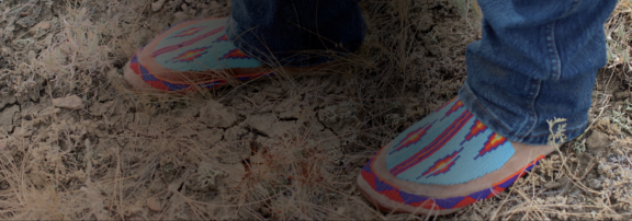 A low-angle shot of a person’s feet wearing colorful, patterned traditional footwear on a dirt path.