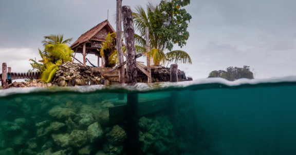 A split-level shot showing a tropical Indigenous hut on stilts above water and the underwater view of coral reefs below.