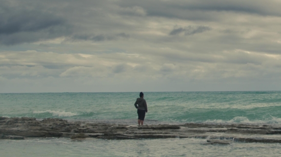 An Indigenous person walking alone along a vast, misty beach at the edge of the tide.