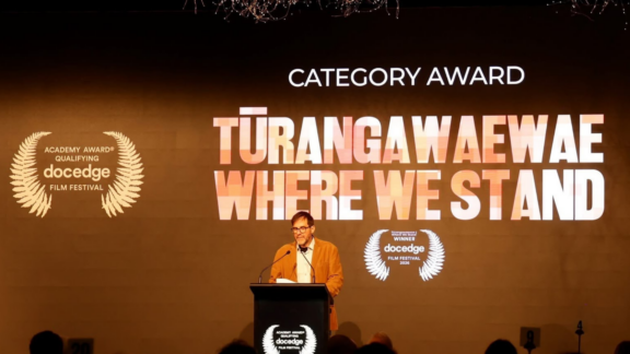 Doc Edge Festival Festival Producer Hunter Weeks stands at a podium to present "Remathau: People of the Ocean" with the "Tūrangawaewae / Where We Stand" Award.