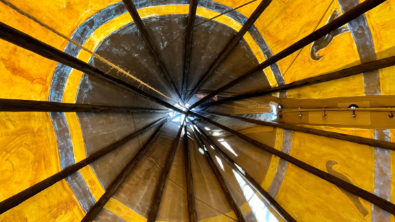 Inside an Indigenous hut, looking up at the central wooden structure draped with yellow fabric.