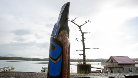 A wooden carving stands along the coastal lands of the Heiltsuk Nation with the ocean in the background.
