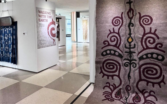 Wide shot of art hanging in the foreground made of felt and wool with other pieces in the background and a wall hanging describing the exhibit.