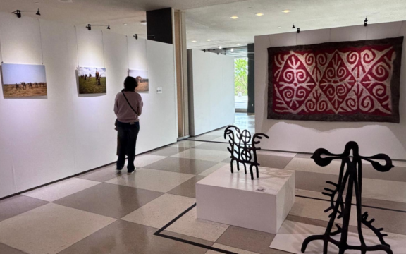 Wide shot of the Origins Fellows "Walking the Earth" exhibit showing sculptures in the foreground and photos and textiles in the background while an attendee reads captions.