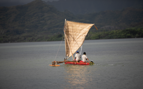 A small sailboat with a large tan sail carrying a few people across a calm water.